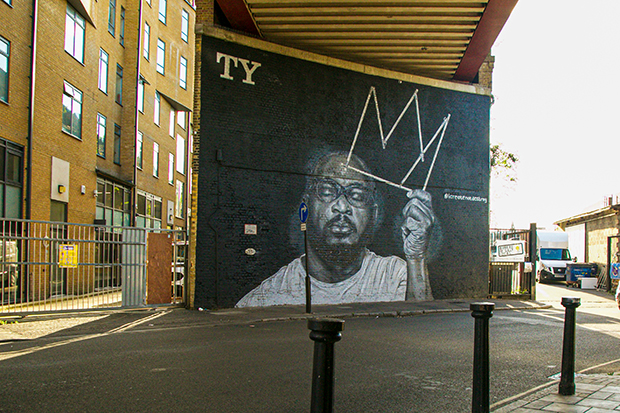 Mural painted on a brick wall at street level, beneath a railway bridge. The mural depicts the rapper TY, who died in 2020. It is a black-and-white painting. TY has his eyes closed and holds the outline of a crown in one hand.
