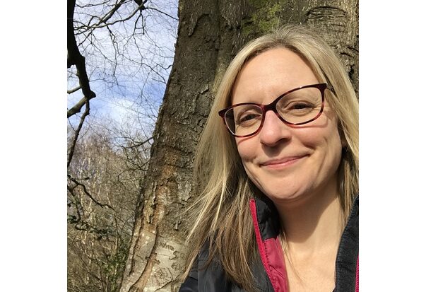Laura is wearing glasses and smiling at the camera. She’s stood in front of a large tree trunk, with more trees and blue sky behind her.