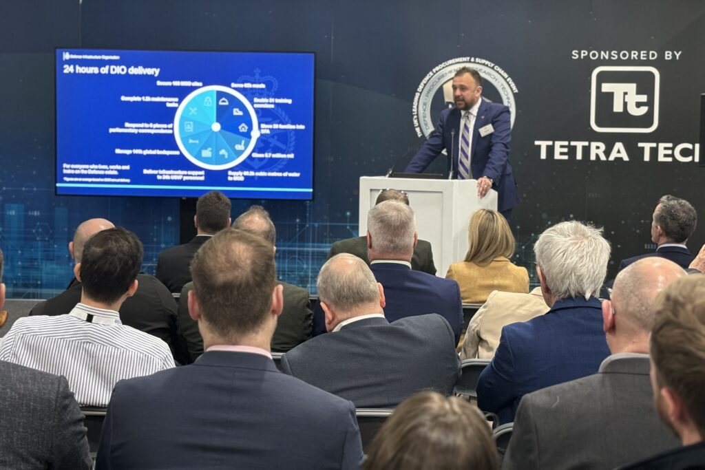 Lee Taylor presenting his speech at the 2024 DPRTE event, to an audience of numerous seated people. He stands behind a lectern and has a screen to his right with a pie chart saying '24 hours of DIO delivery'. The wall behind him reads 'Sponsored by Tetra Tech'.