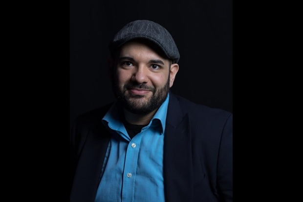 Photograph of a man with a beard, wearing a grey flat cap, blue button-up shirt, and dark blazer against a black background.