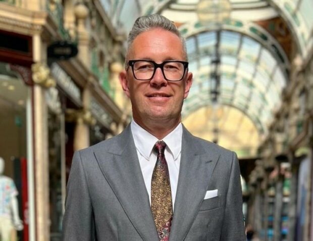 CDIO Richard Corbridge stood in open air shopping centre in suit, tie and glasses. Smiling to camera.