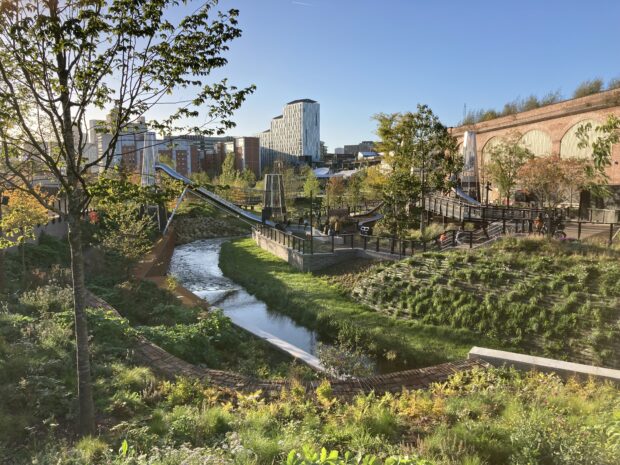 Image shows an urban park with lots of trees and greenery surrounding the buildings There is a canal running through the centre of the image, and tall buildings in the background. A blue sky hangs overhead.