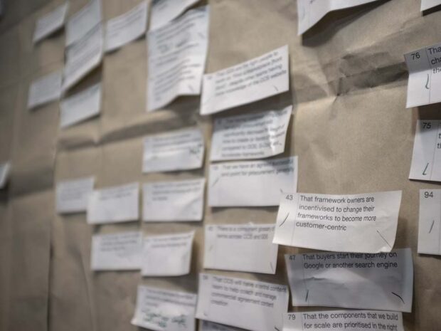 Numerous small pieces of white paper with an assumption typed on each one arranged in columns on a large piece of brown paper on a wall