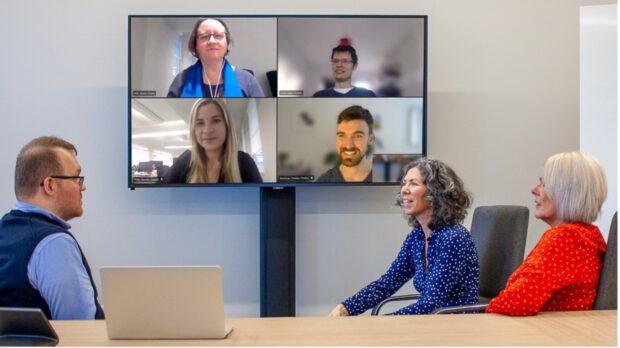 A team meeting with some of the team together in a meeting room and others dialed-in on a screen. 