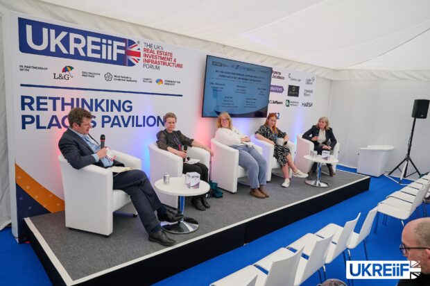 A man and 4 women sit on individual armchairs on a stage, each with a microphone, engaged in a panel discussion in front of an audience.