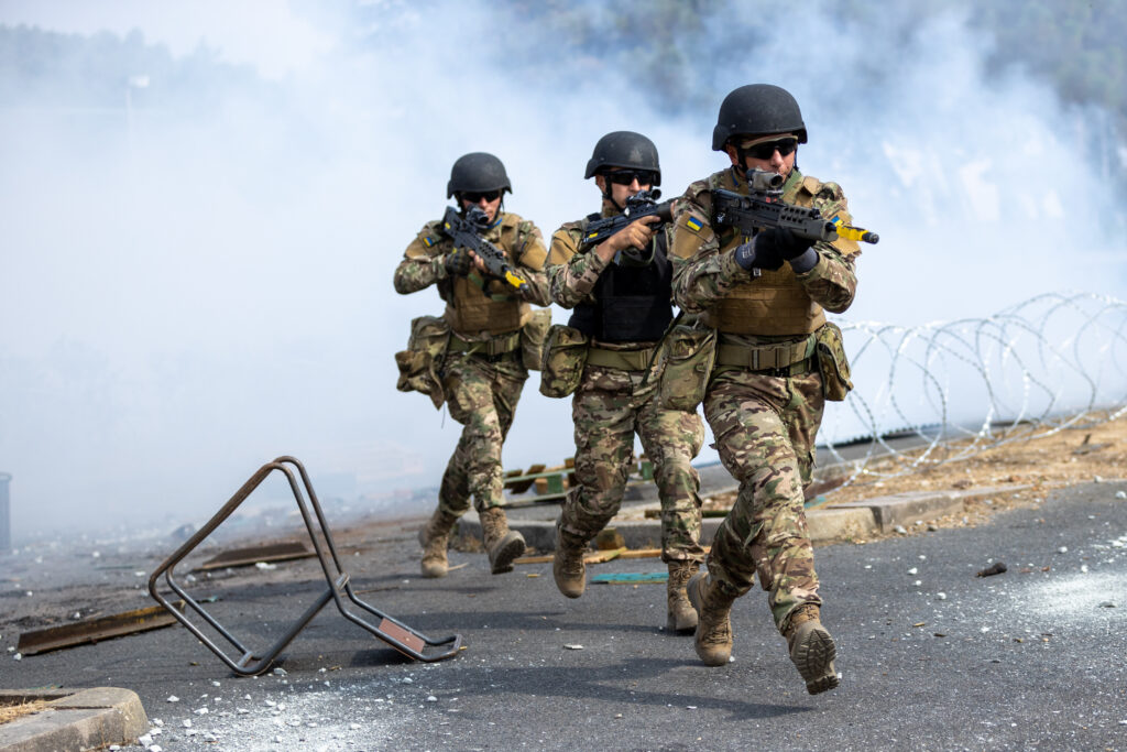 Three Ukrainian soldiers run on a tarmac surface with weapons up and ready. There is barbed wire and an overturned chair on the ground and smoke in the background.