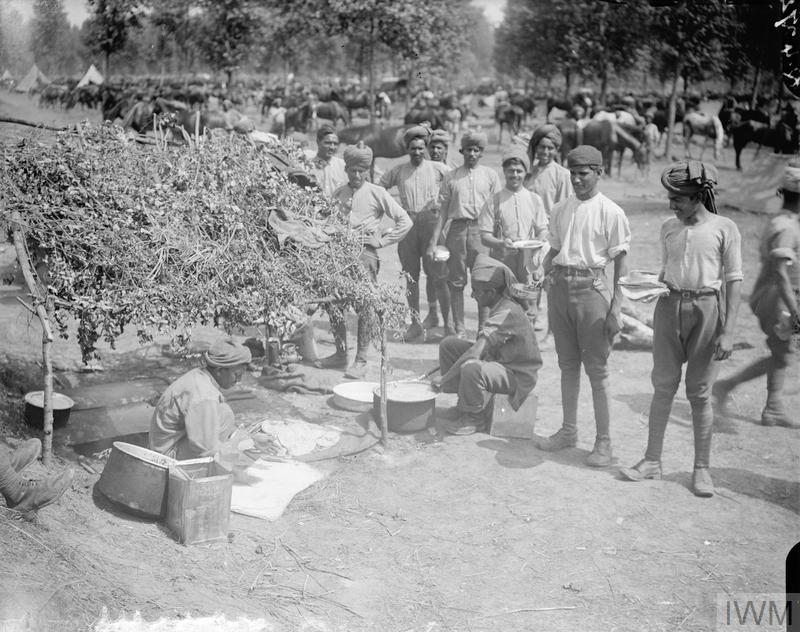A group of Indian soldiers, possibly soldiers of the 9th Hodson's Horse (wearing just shirts and trousers), stand round the improvised cookhouse at an Indian cavalry camp near Querrieu whilst two seated cooks prepare some food. Indian Cavalry Camp. 29 July 1916.