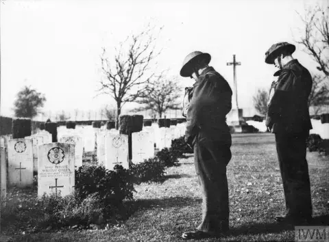 Two soldiers stand with heads bowed in front of a row of war graves. 