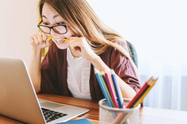 A girl biting into a pencil, looking pleased at something on her computer screen