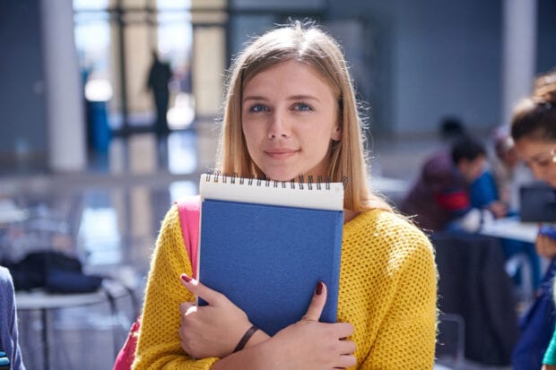 Female student, holding study books, smiles at camera.