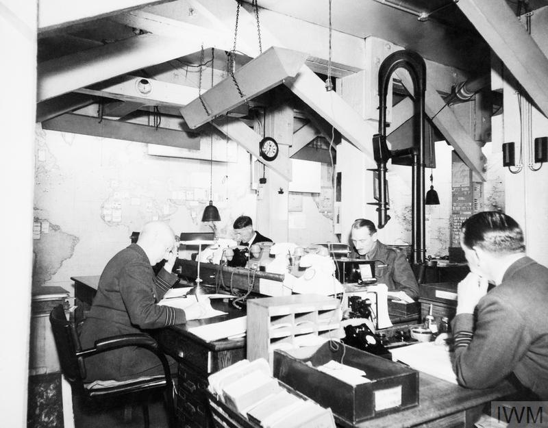 Four military officers sit at desks while working in an underground bunker in 1945. There are maps pinned to the walls behind them. 