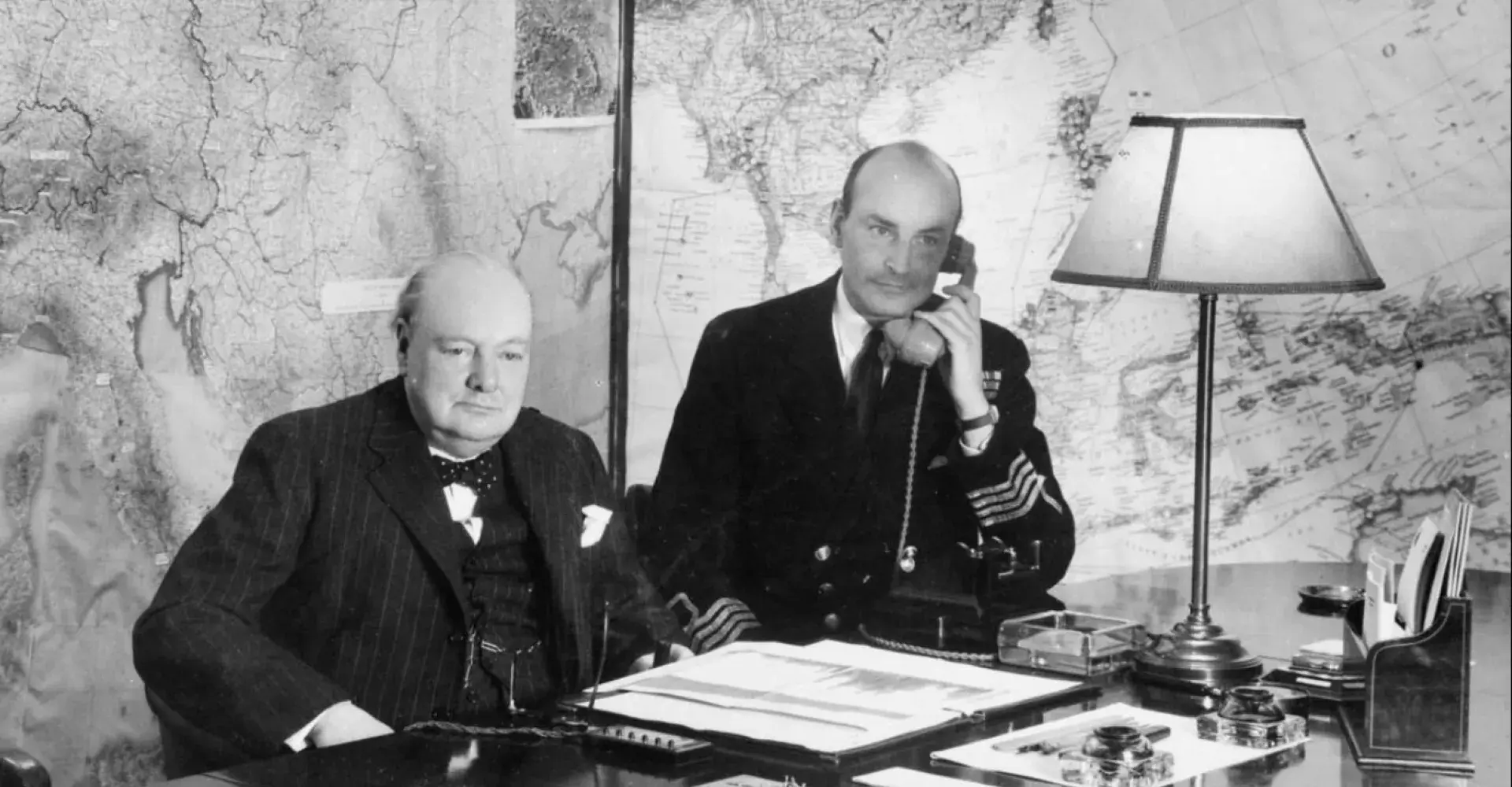 The Prime Minister Winston Churchill sits at his desk in the Map Room at the Cabinet War Rooms. Beside him , Captain Pym of the RNVR takes a telephone call.