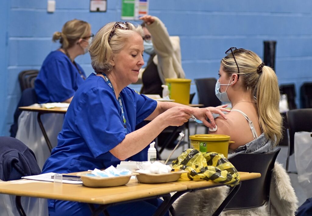 Healthcare worker in blue medical uniform administers a meningitis vaccination to a seated student at a temporary vaccination clinic on a university campus. Medical supplies and sharps containers are visible on tables, with additional healthcare staff and students in the background. The vaccination session took place at Canterbury University in Kent, UK, on 20 March 2026, following a recent meningitis outbreak