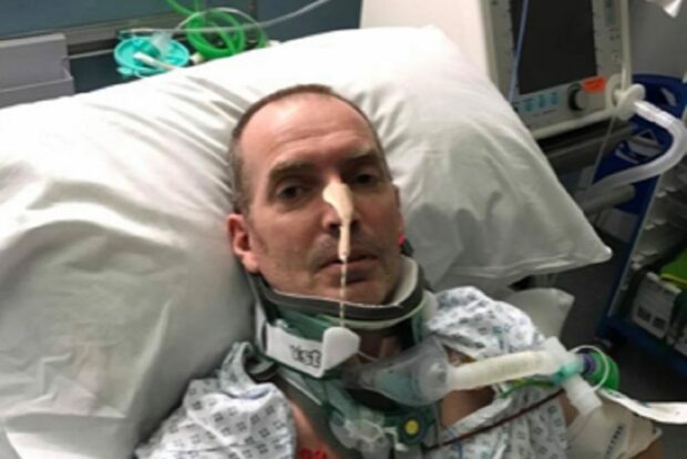 A man is sitting in a hospital bed, with several tubes attached from his body to a monitor.