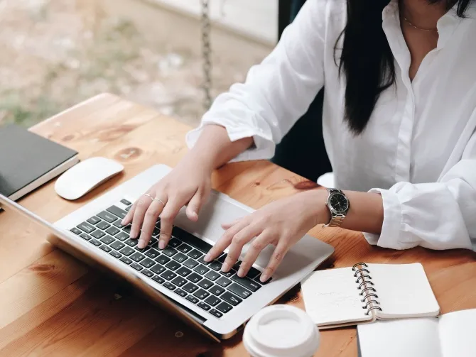View of a young woman's hands typing on a laptop on a modern wooden desk, a notebook open next to the keyboard
