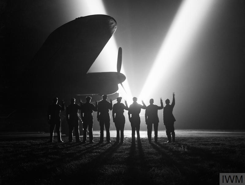 Ground crew on a RAF Bomber Command station in Britain return the V-sign to a neighbouring searchlight crew . Silhouetted is the nose of a Lancaster.