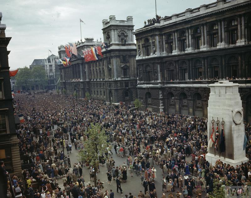 Crowds gather in front of the Ministry of Health building and Cenotaph in Whitehall, 8 May 1945. The balcony and roof of the building are decorated with flags. 