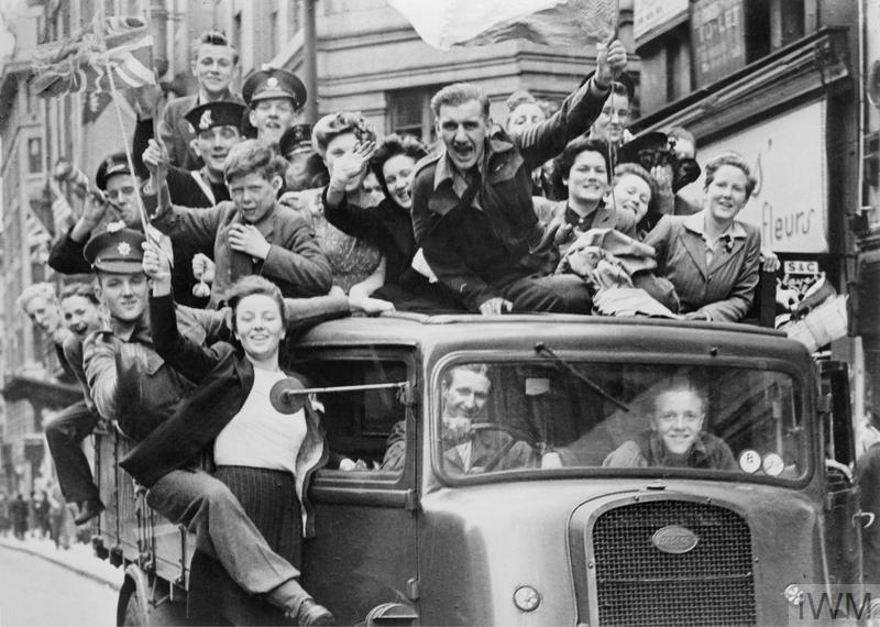 A crowded truck of smiling revellers, some waving flags, pass through the Strand, London, 8 May 1945.