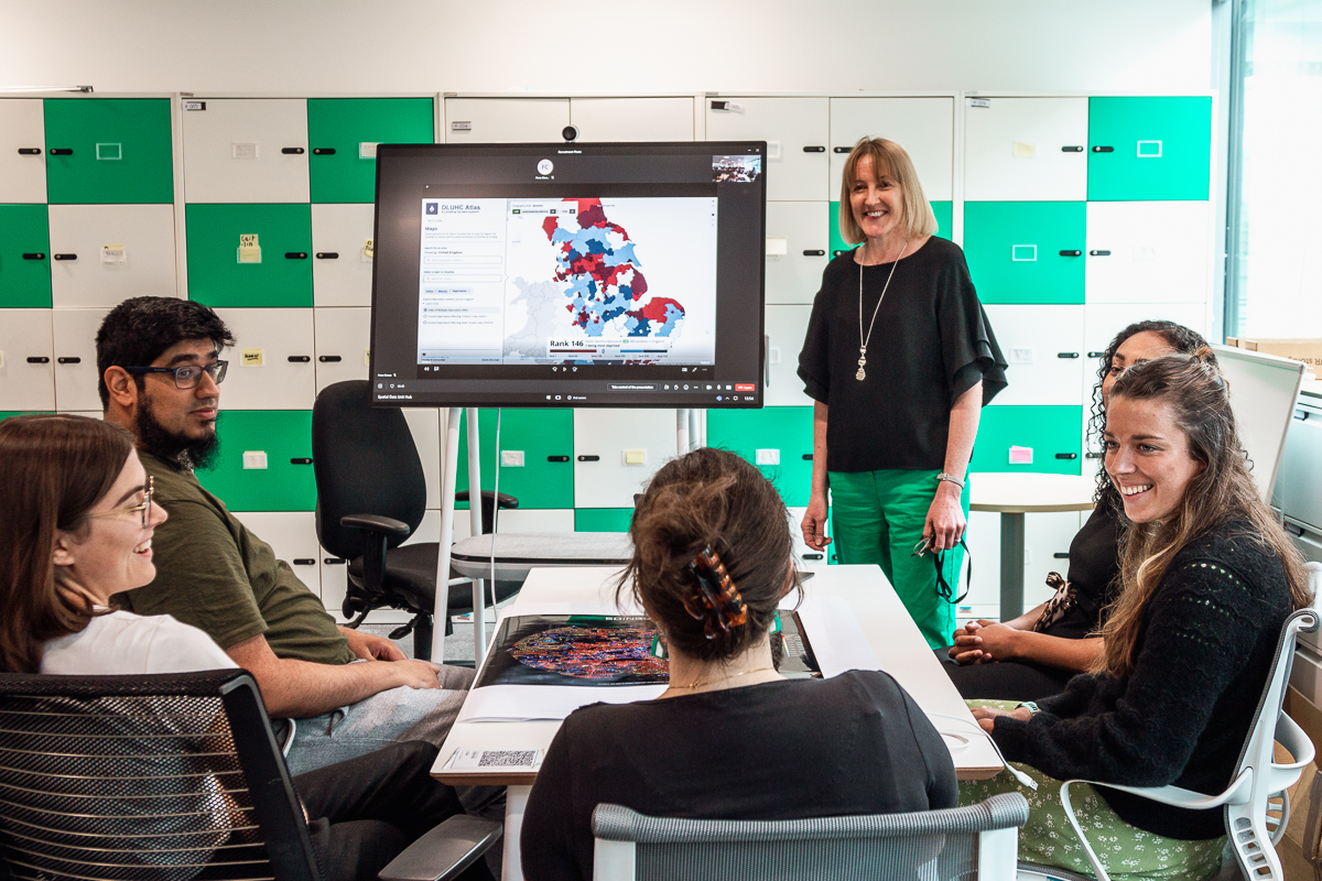 Fiona Clowes standing in front of a monitor which shows a digital map of regions in England. She is surrounded by a group of colleagues who sit at a desk.