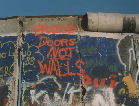 A man chips away at a part of the Berlin Wall. Grafffiti above him reads 'Build Doors Not Walls.' Still from the NATO film collection.
