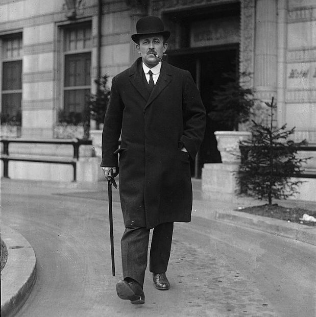 Sir Maurice Hankey is striding with a sense of purpose. He is wearing a bowler hat, smoking a pipe and carrying a walking stick.
