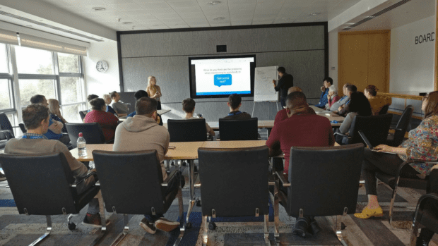 A room of people watching a presentation with one presenter looking at a large screen and the other writing on a piece of paper stuck to a whiteboard.