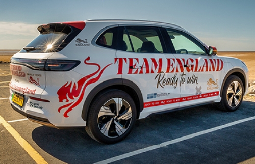 Branded Team England and Sport England electric taxi parked by a wide sandy beach on a sunny day under a clear blue sky.