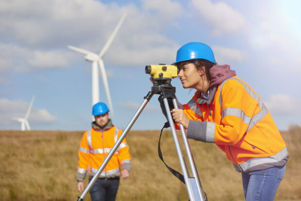 two wind farm engineers using a builder's level to plan out the expansion of the wind farm site. they are wearing orange high visibility jackets and blue hard hats. One is male , one is female. In the foreground the female is looking through the level whilst the male engineer is approaching .In the background wind turbines can be seen across the landscape.