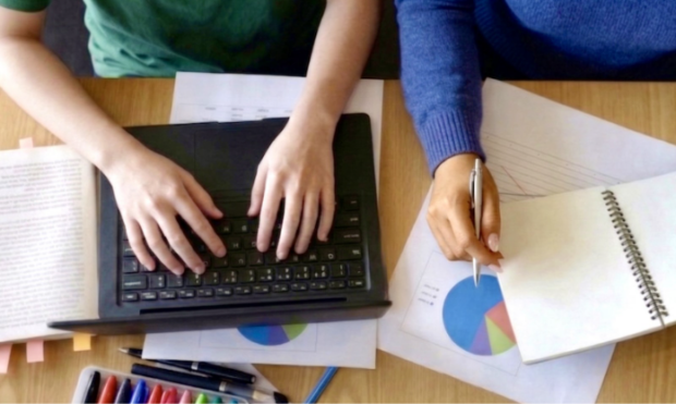 A young person's hands typing on a laptop, with an adult's hand next to it holding a pen
