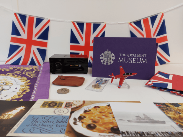 The contents of a 'Museum in a box' with Union Jack bunting and flags, a Jubilee teatowel, recipes from the Silver Jubilee, a picture of a warship, a model airplane, a small red purse and a coin.