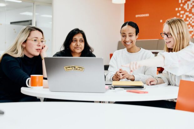 Four colleagues are working together around a laptop, smiling. One of the colleagues is pointing at the screen.