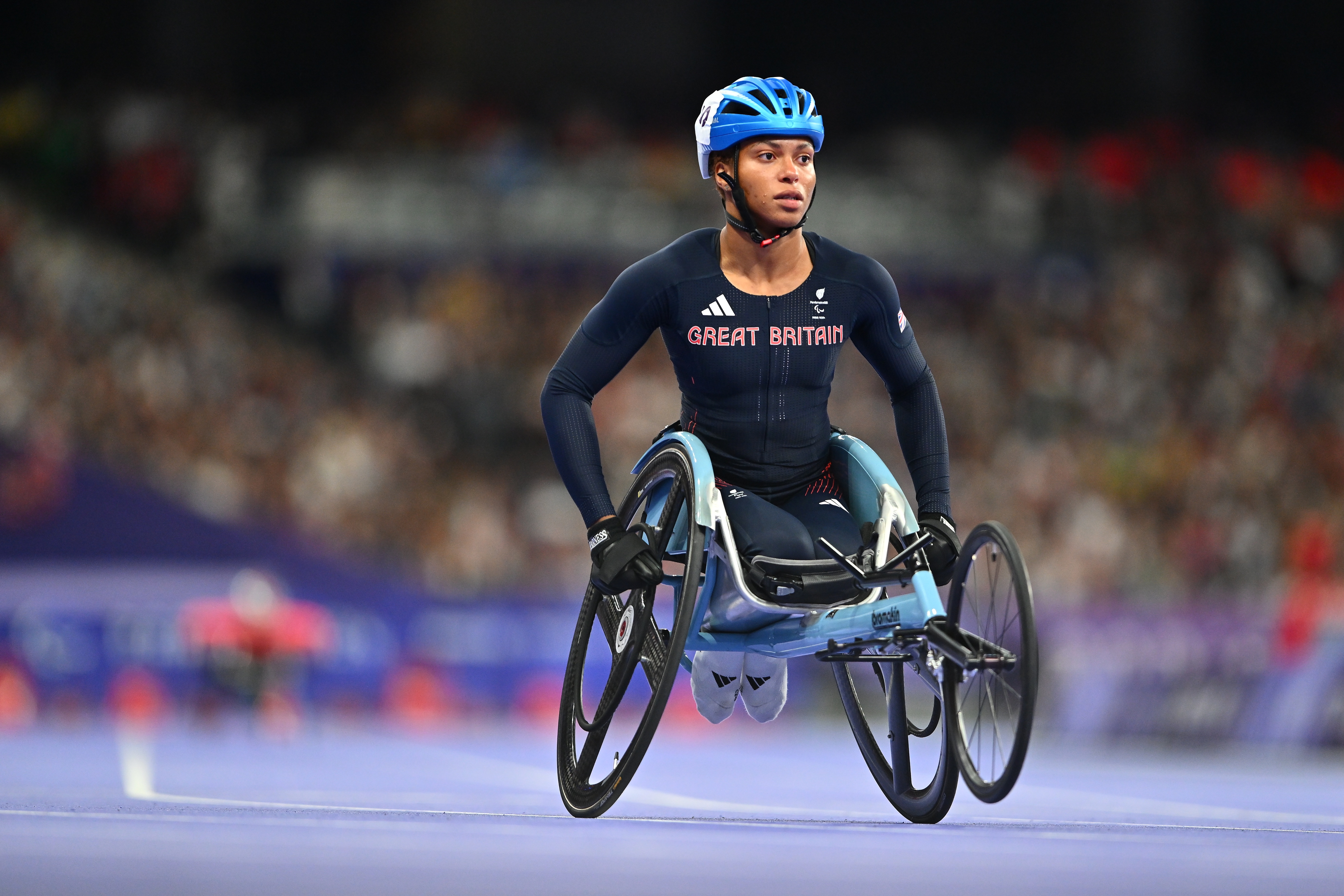 Athlete in a navy Great Britain racing suit and blue helmet competes in a Paralympic track event, propelling a racing wheelchair around an indoor track with a blurred crowd in the background.