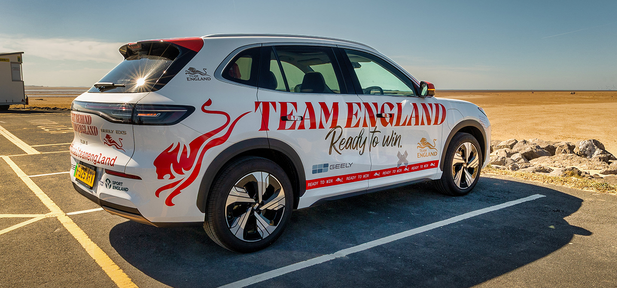 Branded Team England and Sport England electric taxi parked by a wide sandy beach on a sunny day under a clear blue sky.