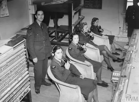 Four women sit in chairs listening to reports through headsets, with two other people standing in the room near them. 