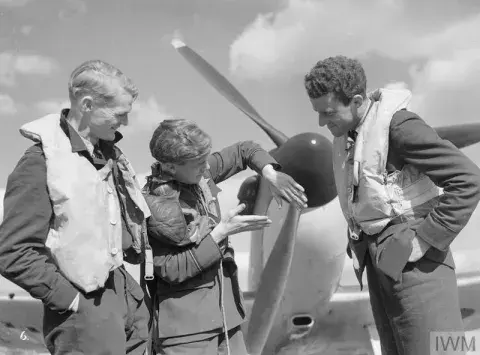 Three pilots dressed in uniform stand in front of an aircraft, having a conversation. 