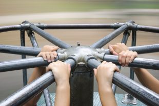The hands of young children's hang on tightly to the bars of a spinning merry go round. There is motion blur in the background, creating a soft wash of brown and green color. There is copy space at the top of the image for a line or two of text.