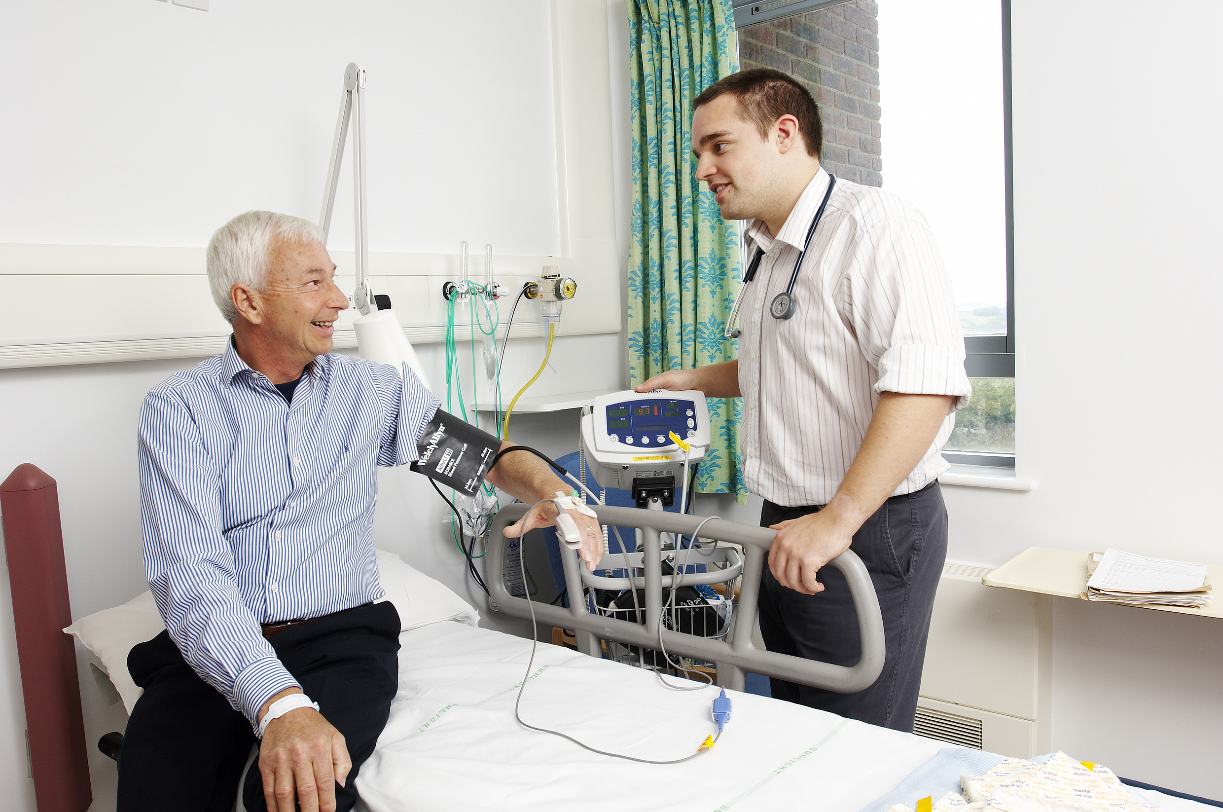 Unique ID: FRAM431 Caption: A doctor measures talks to a patient while taking his blood pressure and heart rate using electronic monitoring equipment in hospital. Assessment. Medical equipment. Restrictions: NHS Photo Library - for use in NHS, local authority Social Care services and Department of Health material only Copyright: ©Crown Copyright