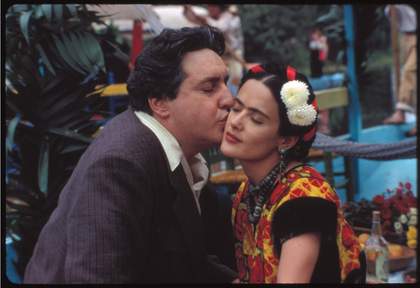 A man in a suit kisses a woman with flowers in her hair on the cheek. The woman wears a colourful dress and sits with her eyes closed outdoors.