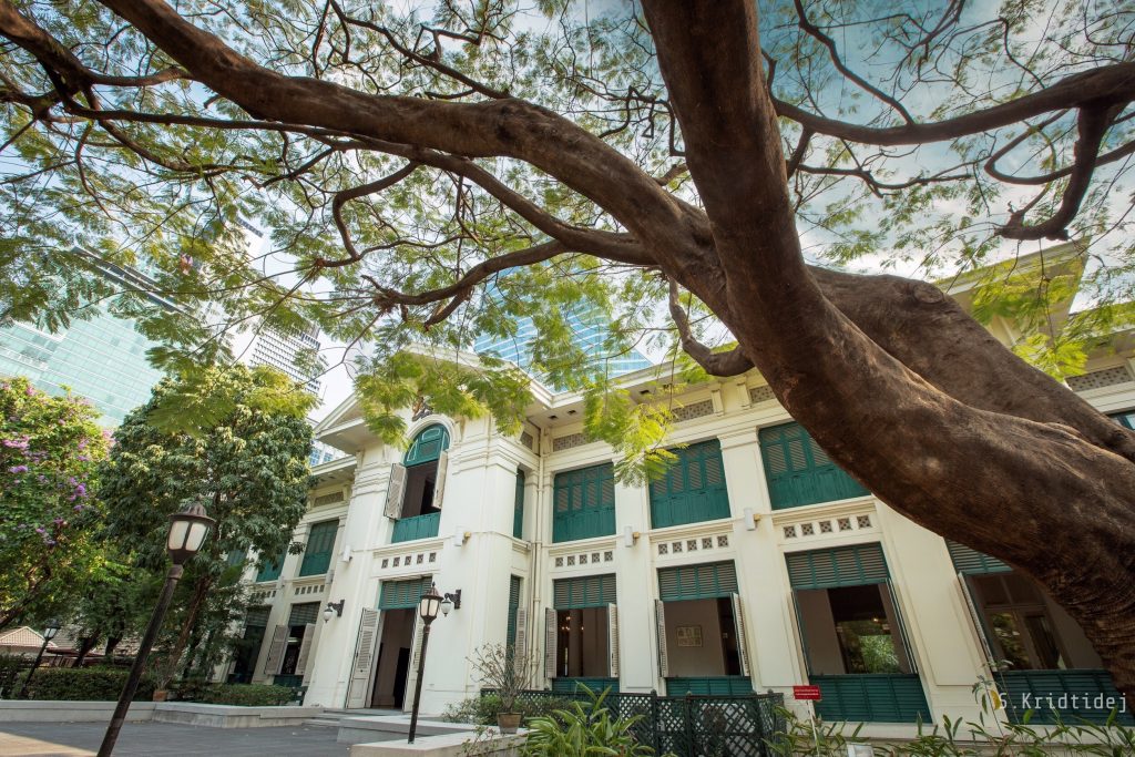 The frontage of the British Embassy Bangkok, March 2019. There is a far spreading tree in the foreground and the Embassy building is an imposing structure - it is mostly white with green shutters.