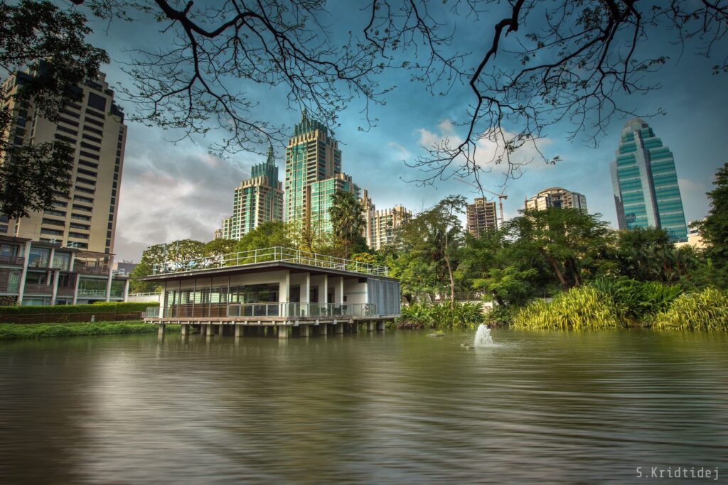 The Nai Lert pond, 2019. A small viewing structure, raised on pillars sits within the pond. A small fountain, within the pond, is also shown. Tall modern buildings dominate the skyline.