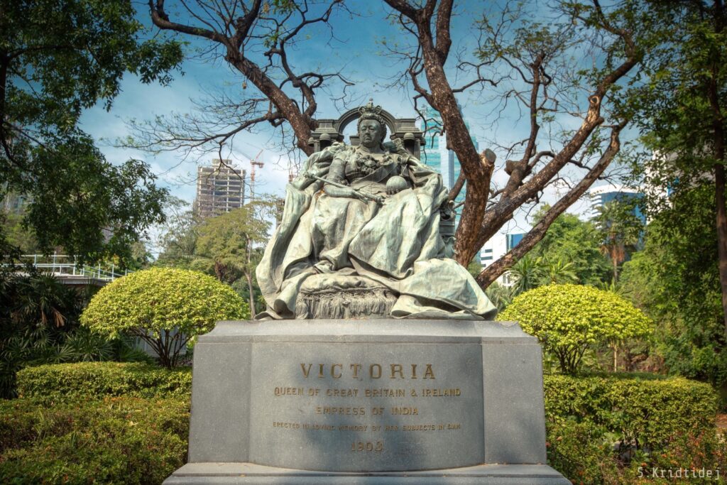 Queen Victoria Memorial in the compound of the British Embassy Bangkok, 2019. The sculpture on the plinth shows Queen Victoria sitting on the throne holding the orb and sceptre. The words on the plinth read: “Victoria, Queen of Great Britain & Ireland, Empress of India. Erected in loving memory by her subjects in Siam, 1903". The gardens are shown in the background, including examples of topiary.