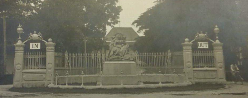 The Queen Victoria Memorial at the British Embassy Bangkok. The central scuplture shows Queen Victoria sat on the throne. Behind it is a crescent shaped structure made of stone with railings with an 'In' sign attached to arches on the left and an 'Out' sign attached to arches on the right