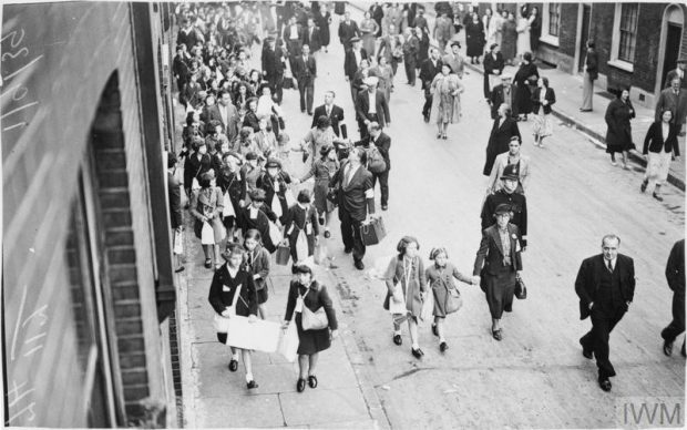 An early start to evacuation is made by children of Myrdle School in Stepney. The children assembled at school at 5am on Friday 1 September 1939. Evacuee children and adults are shown walking along a street carrying suitcases and gas mask boxes. Some of the adults are wearing arm bands which identify them as volunteer marshals.