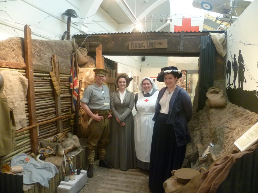 A photograph of two re-enactors dressed as a Tommy and a VAD nurse and two members of the museum staff dressed in Edwardian costume. The photograph is taken in the trench exhibit which is part of the exhibition.