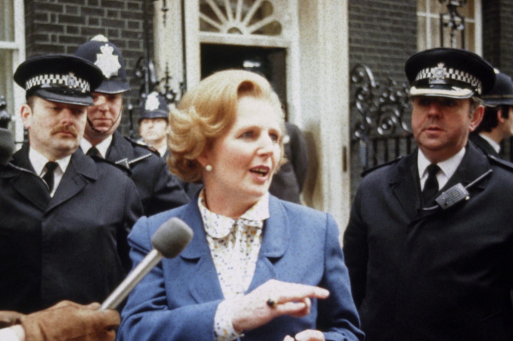 Margaret Thatcher arrives at Number 10 Downing Street in 1979. She is flanked by police officers and speaking to a microphone.