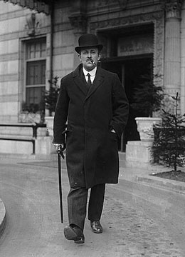Maurice Hankey is striding with a sense of purpose. He is wearing a bowler hat, smoking a pipe, and carrying a walking stick.