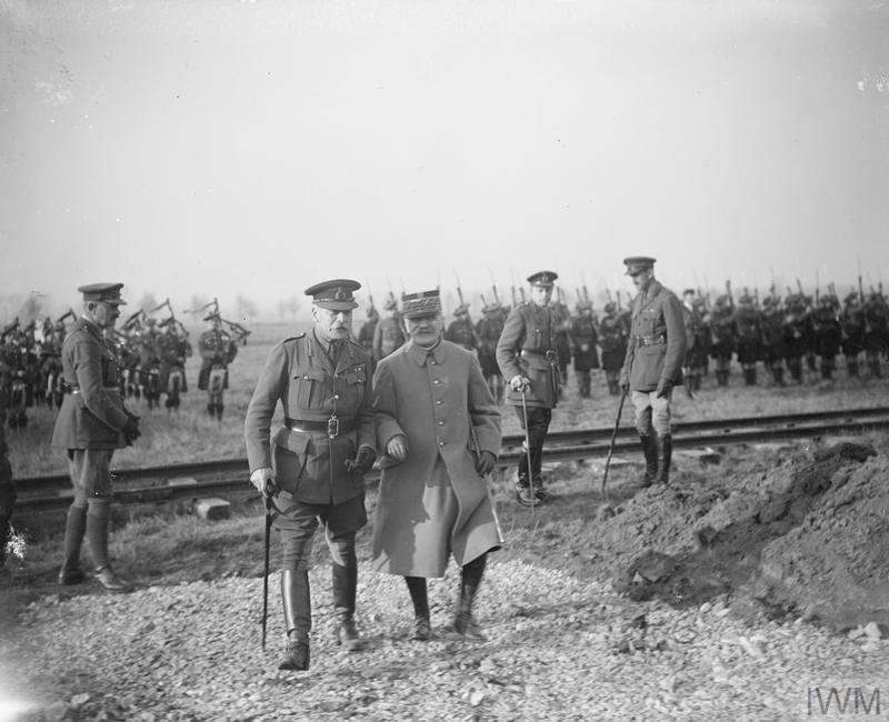 The picture displays 2 men walking and discussing, looking like they discussing what is in front of them. They are surrounded by younger soldiers in formation.