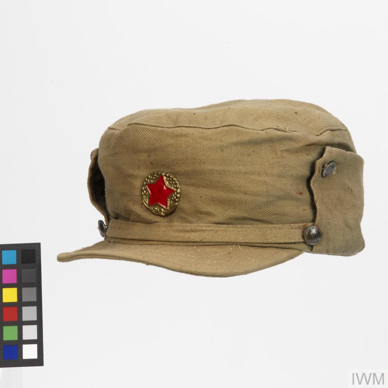 Non-rigid field cap of green fabric. The cap features a cloth chinstrap and a five-pointed red star on a gold circular metal badge attached to the front.