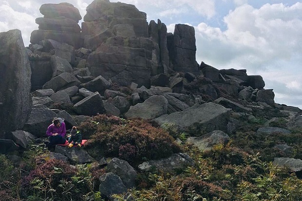A sunny day in the Peak District. There are tall rocks in the background with lots of boulders, moss and ferns in front of them. Jeni and a toddler are sat on a blanket on a rock near the front, wearing coats and hats.