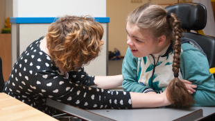 A young girl is sitting in her wheelchair, communicating with a teacher who is leaning forward to talk to her.